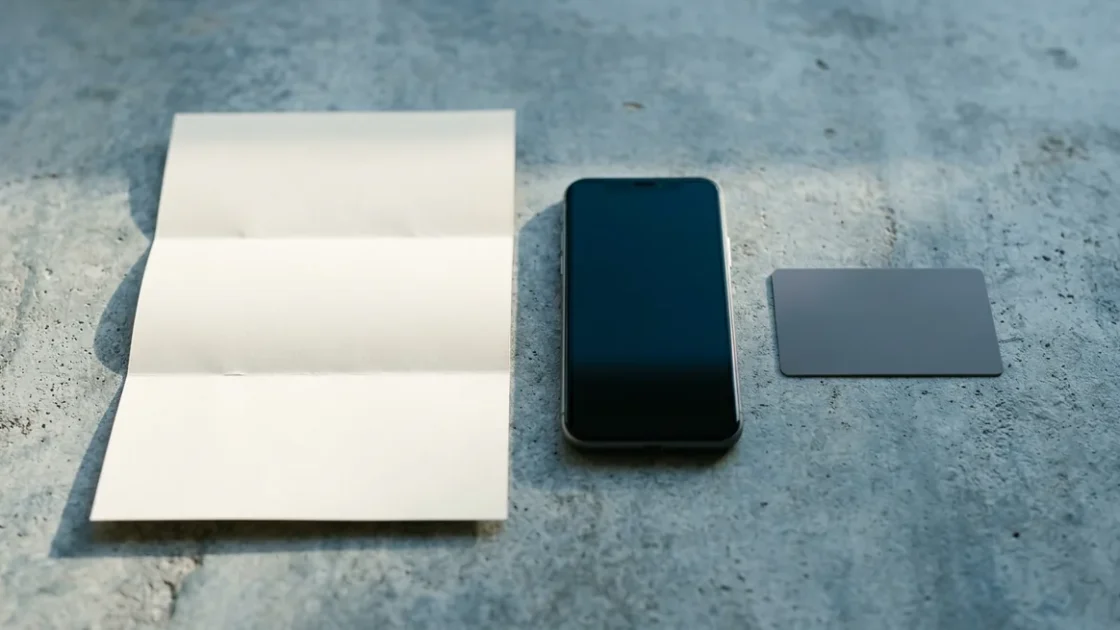Aqui estão algumas opções curtas e descritivas: 1. **Documento dobrado, celular de tela escura e cartão em branco sobre concreto minimalista, iluminados por forte luz lateral.** 2. **Objetos em bra